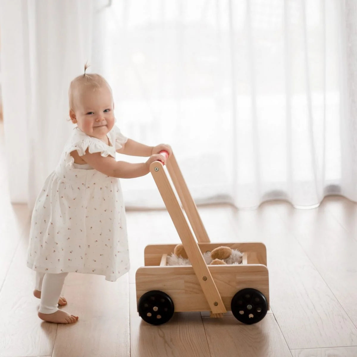 Child in a white dress pushing a wooden toy cart in a bright room.
